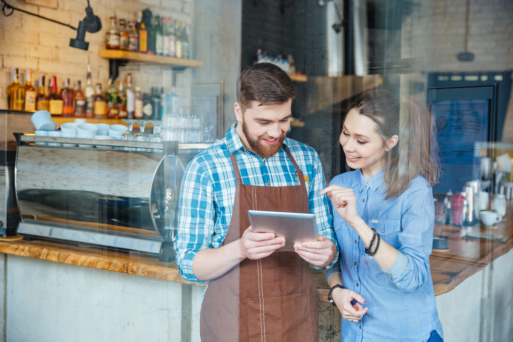 Smiling handsome waiter holding tablet and young pretty woman pointing on it in coffee shop